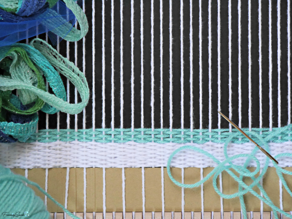 Weaving project in progress with small frame loom. White cotton warp threads are wrapped. Weaving with turquoise yarn has begun. @FanningSparks