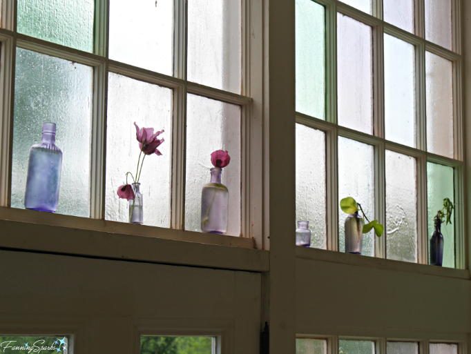 Simple colored glass bottles displayed with a few choice blooms echo the colored glass window panes. @FanningSparks